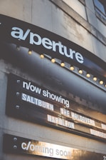 A marquee sign displays movie titles under a theater sign labeled 'aperture'. The current showings include 'Saltburn', 'The Holdovers', and 'Napoleon', with a section noting upcoming features. The structure is illuminated by small round lights, and the signage appears on a building's exterior.
