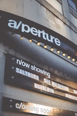 A marquee sign displays movie titles under a theater sign labeled 'aperture'. The current showings include 'Saltburn', 'The Holdovers', and 'Napoleon', with a section noting upcoming features. The structure is illuminated by small round lights, and the signage appears on a building's exterior.