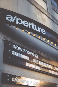 A marquee sign displays movie titles under a theater sign labeled 'aperture'. The current showings include 'Saltburn', 'The Holdovers', and 'Napoleon', with a section noting upcoming features. The structure is illuminated by small round lights, and the signage appears on a building's exterior.