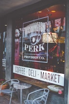 A storefront window displays the signage for 'Washington Perk and Provision Co.', featuring a graphic of a coffee cup. The interior appears warmly lit with hanging lights and decorative elements inside. Outside, there is a small seating area with two metal chairs and a round table.