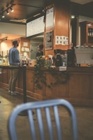A cozy coffee shop interior with wooden furnishings and warm lighting. A man in casual attire stands at the counter speaking with a barista. The counter is adorned with coffee cups, a cash register, and a menu board. There is a plant adding a touch of greenery. In the foreground, a blurred chair partially obstructs the view.