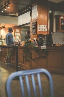 A cozy coffee shop interior with wooden furnishings and warm lighting. A man in casual attire stands at the counter speaking with a barista. The counter is adorned with coffee cups, a cash register, and a menu board. There is a plant adding a touch of greenery. In the foreground, a blurred chair partially obstructs the view.