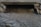 A concrete drainage culvert partially submerged in flowing water, surrounded by rocks and some fallen leaves. The surface of the water is rippling, and the interior of the culvert is dark.