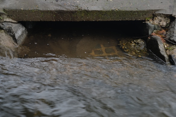A concrete drainage culvert partially submerged in flowing water, surrounded by rocks and some fallen leaves. The surface of the water is rippling, and the interior of the culvert is dark.