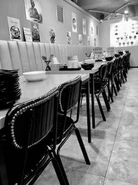 A restaurant interior with a long row of empty tables and chairs, neatly aligned along a tiled floor. The tables are set with various condiments, chopsticks, and stacks of bowls and plates. The walls are decorated with food posters, and the lighting is provided by overhead lamps, creating a clean and organized dining atmosphere.