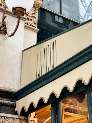 An elegant storefront with intricate architectural detailing, featuring a decorative lamp and a stylish sign with the word 'Cracco'. The building materials include ornate stonework and glass.