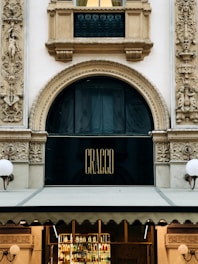 The image features an ornate architectural facade with intricate carvings and details surrounding a large arched window. Above the window, there is a black sign with the word 'CRACCO' in gold letters. Below, the entrance appears to display various bottles, suggesting a restaurant or bar setting. Flanking either side of the facade are decorative stone reliefs and lamps.