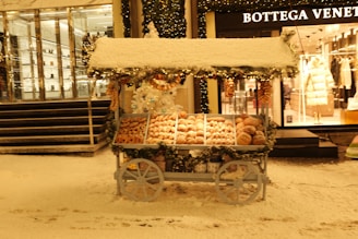 A cart laden with assorted pastries and doughnuts stands on a snowy street next to a luxury store. The cart is adorned with festive decorations, including garlands and wreaths. Snow covers the ground and the top of the cart, creating a wintry atmosphere. In the background, elegant shop windows display stylish clothing.
