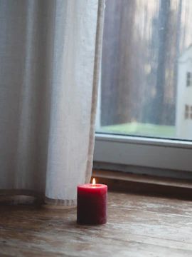 Cozy home setting with lit burgundy and gold candles on a wooden table near a window.