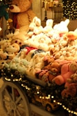 A festive display of plush toys is arranged on a cart, featuring a variety of stuffed animals including teddy bears, bunnies, and other cuddly creatures. The cart is adorned with decorative garland and lights, enhancing the warm and cozy ambiance. Behind the display, a large teddy bear looms over the collection, contributing to the inviting and whimsical atmosphere.