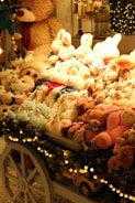 A festive display of plush toys is arranged on a cart, featuring a variety of stuffed animals including teddy bears, bunnies, and other cuddly creatures. The cart is adorned with decorative garland and lights, enhancing the warm and cozy ambiance. Behind the display, a large teddy bear looms over the collection, contributing to the inviting and whimsical atmosphere.