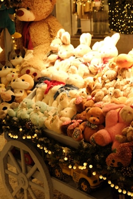 A festive display of plush toys is arranged on a cart, featuring a variety of stuffed animals including teddy bears, bunnies, and other cuddly creatures. The cart is adorned with decorative garland and lights, enhancing the warm and cozy ambiance. Behind the display, a large teddy bear looms over the collection, contributing to the inviting and whimsical atmosphere.