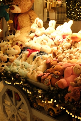A festive display of plush toys is arranged on a cart, featuring a variety of stuffed animals including teddy bears, bunnies, and other cuddly creatures. The cart is adorned with decorative garland and lights, enhancing the warm and cozy ambiance. Behind the display, a large teddy bear looms over the collection, contributing to the inviting and whimsical atmosphere.