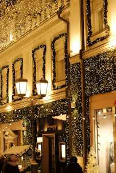 A building facade is decorated with numerous small golden lights and green garlands, suggesting holiday or festive decor. Two large street lamps are illuminated, adding to the warm glow. People are visible on the street, and shop windows and doorways are adorned with decorations.