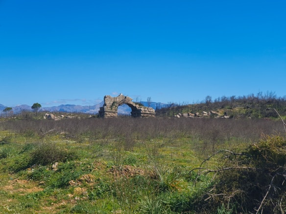 A vast open landscape featuring a partially ruined stone arch standing solitary in a grassy field. Sparse vegetation covers the ground, and distant arid hills or mountains are visible under a clear, blue sky.