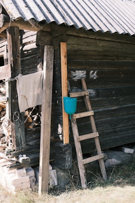 A weathered wooden cabin with a tin roof and rustic ladder leaning against its side. A blue water container is attached to one of the wooden planks. Nearby, a pile of cut wood is partially covered with a cloth.