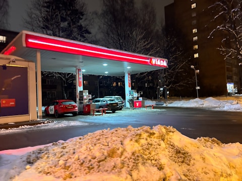 A gas station with a bright red and white canopy is lit up at night. Snow covers the ground, and several cars are parked by the gas pumps. Trees and apartment buildings are visible in the background, with soft lighting from the surrounding street lamps.
