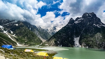 A breathtaking landscape of a serene lake surrounded by snow-capped mountains under a partly cloudy sky. Vibrant blue and yellow tents are set up on the green pasture near the lake, suggesting a camping site. The mountains tower majestically, creating a contrast with the tranquil waters below.