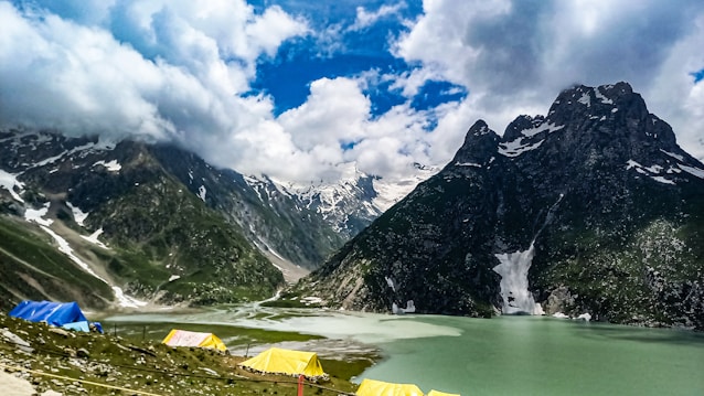 A breathtaking landscape of a serene lake surrounded by snow-capped mountains under a partly cloudy sky. Vibrant blue and yellow tents are set up on the green pasture near the lake, suggesting a camping site. The mountains tower majestically, creating a contrast with the tranquil waters below.