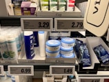 A supermarket shelf displays a variety of skincare and personal care products. On the left, there are white bottles labeled Satin Care with aloe vera graphics. In the middle, blue and white bottles of Cien branded body lotions and creams are organized in boxed displays with clearly visible price tags above them. To the right, razor packs are neatly arranged, also marked with the Cien brand. The shelf is well-organized with different product categories neatly segregated.