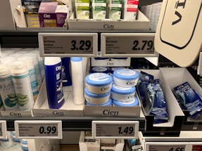 A supermarket shelf displays a variety of skincare and personal care products. On the left, there are white bottles labeled Satin Care with aloe vera graphics. In the middle, blue and white bottles of Cien branded body lotions and creams are organized in boxed displays with clearly visible price tags above them. To the right, razor packs are neatly arranged, also marked with the Cien brand. The shelf is well-organized with different product categories neatly segregated.