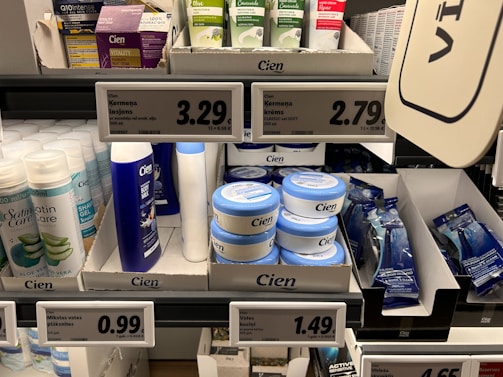 A supermarket shelf displays a variety of skincare and personal care products. On the left, there are white bottles labeled Satin Care with aloe vera graphics. In the middle, blue and white bottles of Cien branded body lotions and creams are organized in boxed displays with clearly visible price tags above them. To the right, razor packs are neatly arranged, also marked with the Cien brand. The shelf is well-organized with different product categories neatly segregated.