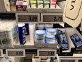 A supermarket shelf displays a variety of skincare and personal care products. On the left, there are white bottles labeled Satin Care with aloe vera graphics. In the middle, blue and white bottles of Cien branded body lotions and creams are organized in boxed displays with clearly visible price tags above them. To the right, razor packs are neatly arranged, also marked with the Cien brand. The shelf is well-organized with different product categories neatly segregated.