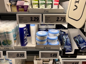 A supermarket shelf displays a variety of skincare and personal care products. On the left, there are white bottles labeled Satin Care with aloe vera graphics. In the middle, blue and white bottles of Cien branded body lotions and creams are organized in boxed displays with clearly visible price tags above them. To the right, razor packs are neatly arranged, also marked with the Cien brand. The shelf is well-organized with different product categories neatly segregated.