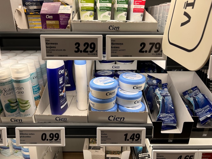 A supermarket shelf displays a variety of skincare and personal care products. On the left, there are white bottles labeled Satin Care with aloe vera graphics. In the middle, blue and white bottles of Cien branded body lotions and creams are organized in boxed displays with clearly visible price tags above them. To the right, razor packs are neatly arranged, also marked with the Cien brand. The shelf is well-organized with different product categories neatly segregated.