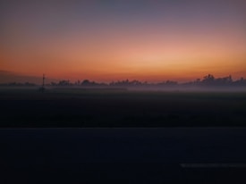 A serene landscape depicting a sunrise or sunset with a gradient of orange and blue hues in the sky. Silhouettes of trees line the horizon, and a mist blankets the ground, creating a mysterious and tranquil atmosphere.