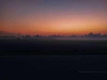 A serene landscape depicting a sunrise or sunset with a gradient of orange and blue hues in the sky. Silhouettes of trees line the horizon, and a mist blankets the ground, creating a mysterious and tranquil atmosphere.