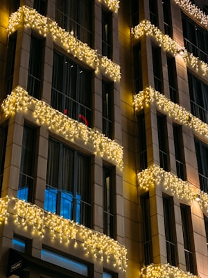 A building facade adorned with strings of warm white lights draped along the balconies. The lights create a festive and inviting atmosphere against the backdrop of the building's modern architecture. Large windows reflect a soft blue hue, adding contrast to the warm lighting.