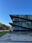 A modern architectural building with sharp, angular features and a textured green facade stands prominently against a clear blue sky. The building's design includes large, dark horizontal elements that intersect with glass panes. Surrounding the structure are well-maintained greenery and trees, along with a concrete pathway leading to the entrance.