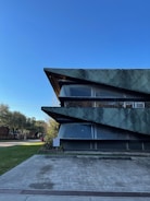 A modern architectural building with sharp, angular features and a textured green facade stands prominently against a clear blue sky. The building's design includes large, dark horizontal elements that intersect with glass panes. Surrounding the structure are well-maintained greenery and trees, along with a concrete pathway leading to the entrance.