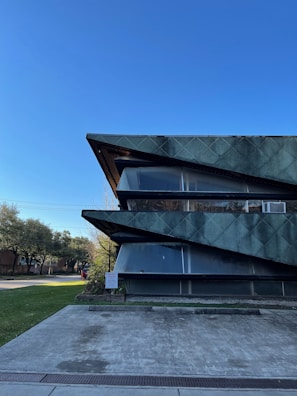 A modern architectural building with sharp, angular features and a textured green facade stands prominently against a clear blue sky. The building's design includes large, dark horizontal elements that intersect with glass panes. Surrounding the structure are well-maintained greenery and trees, along with a concrete pathway leading to the entrance.