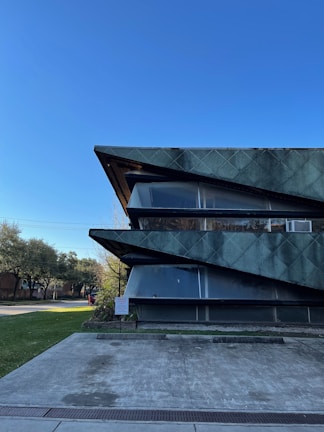 A modern architectural building with sharp, angular features and a textured green facade stands prominently against a clear blue sky. The building's design includes large, dark horizontal elements that intersect with glass panes. Surrounding the structure are well-maintained greenery and trees, along with a concrete pathway leading to the entrance.