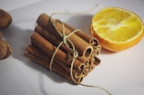 A bundle of cinnamon sticks tied with a string is placed next to a halved orange on a light-colored surface. The arrangement creates a contrast between the rich brown tones of the cinnamon and the vibrant yellow of the orange flesh.