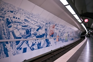 A subway station with a large mural depicting an artistic aerial view of a cityscape using blue and red tones. The mural is displayed along the curved wall adjacent to the train tracks. The station is modern, with sleek design elements, including overhead lighting and a sign indicating 'Rathaus'. The platform is clean and mostly empty, indicating an off-peak time.