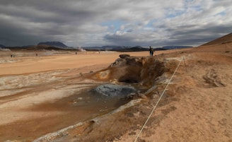 A barren landscape featuring a geothermal area with steam rising from the ground. The terrain is rocky and sandy with a predominant brown and ochre hue. In the background, mountains can be seen under a cloudy sky. A small group of people is exploring the area, and some ropes guide the path around the geothermal formations.