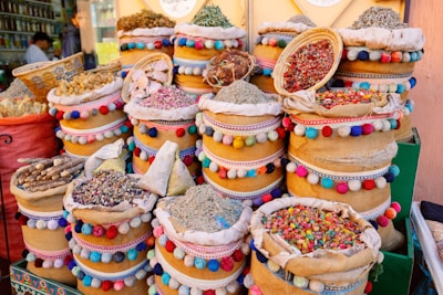 A vibrant display of various dried herbs, spices, and colorful items are arranged in ornate, multi-colored baskets. The baskets are decorated with pom-poms and intricate patterns, adding a lively and cultural touch. The scene suggests a traditional market setting.
