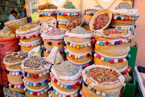 A vibrant display of various dried herbs, spices, and colorful items are arranged in ornate, multi-colored baskets. The baskets are decorated with pom-poms and intricate patterns, adding a lively and cultural touch. The scene suggests a traditional market setting.