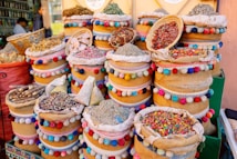 A vibrant display of various dried herbs, spices, and colorful items are arranged in ornate, multi-colored baskets. The baskets are decorated with pom-poms and intricate patterns, adding a lively and cultural touch. The scene suggests a traditional market setting.