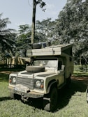 A rugged, off-road vehicle is parked on a grassy area surrounded by lush greenery and tall trees. The vehicle has a spare tire mounted on its hood and another on the roof. The setting is outdoors, and the environment suggests it is in a forest or jungle.