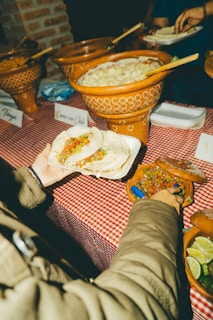 A person is holding two tacos filled with vegetables over a table covered with a red and white checkered tablecloth. Next to the person, there are clay pots with various food dishes, including one labeled as 'Carne con Chile.' Slices of lime and a salsa bowl are visible on the table. Another person in the background appears to be serving food onto a plate.