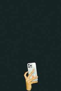 A hand holding a smartphone, which has a colorful sticker on its back. The background is a solid dark color, emphasizing the focus on the hand and phone.