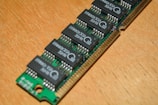A close-up view of a green circuit board featuring several black integrated circuits arranged in a row. The board is placed on a light wooden surface, showing detailed connectors and circuitry.