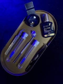 A gold tray holds various tools including a magnifying glass, a small bottle labeled 'Absolute Fast', metal tweezers, two test tubes with brushes inside, a metal scraper, and a dental mirror. A soft blue light casts over the arrangement, creating a subtle glow on the objects.