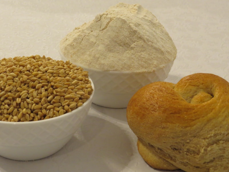 Three white bowls are arranged on a light-colored surface. One bowl contains wheat grains, another bowl is filled with flour, and the third bowl has a freshly baked bun.