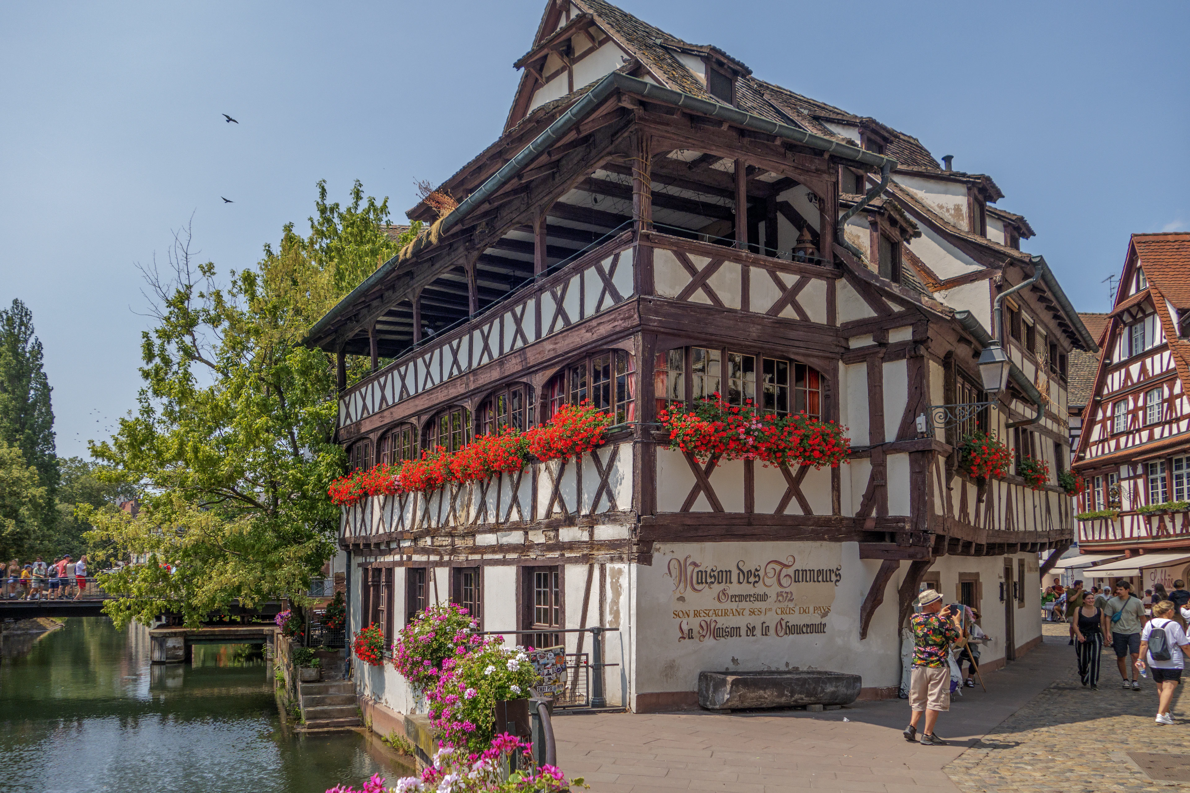 A flower-draped half-timbered building leans over a canal in Strasbourg's Petite France quarter under blue sky.