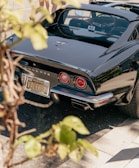 A classic black Corvette sports car is parked, with sunlight reflecting off its glossy surface. The car has distinctive red circular taillights and a California license plate with the name 'VALEREY'. There are some leaves and branches in the foreground, creating a natural frame around the image.
